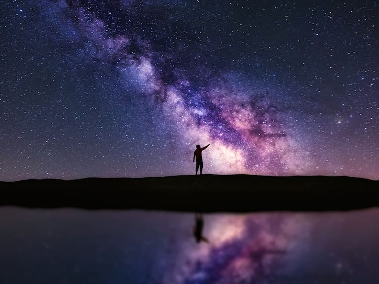 Person pointing up the Milky Way galaxy at night. The scene is reflected in water. A Veiga, Ourense, Spain