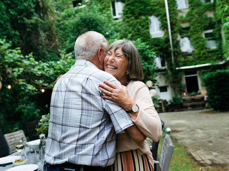 An elderly couple embracing and smiling before sitting down for a barbecue with the family in a courtyard.