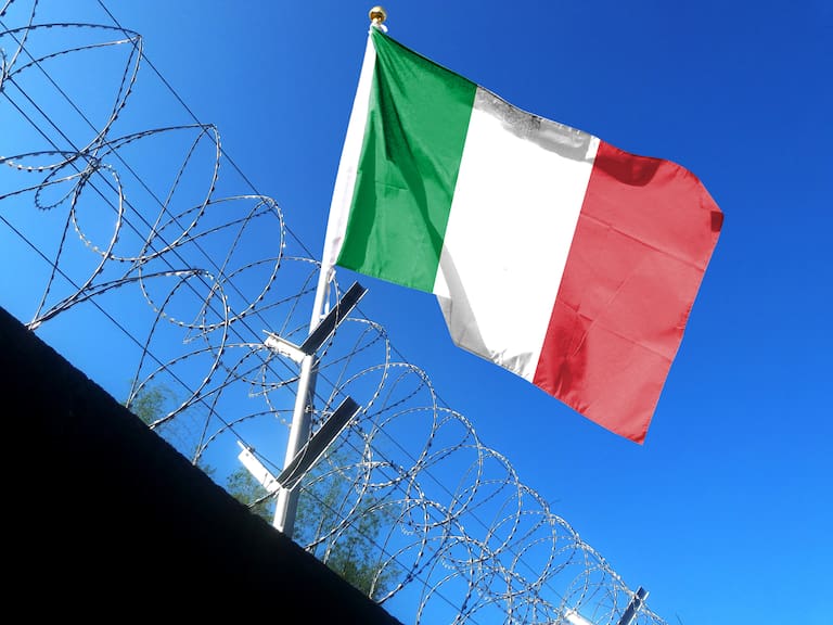 The Italian flag hangs in the cloudy sky outside the prison&#039;s barbed wire. waving in the sky